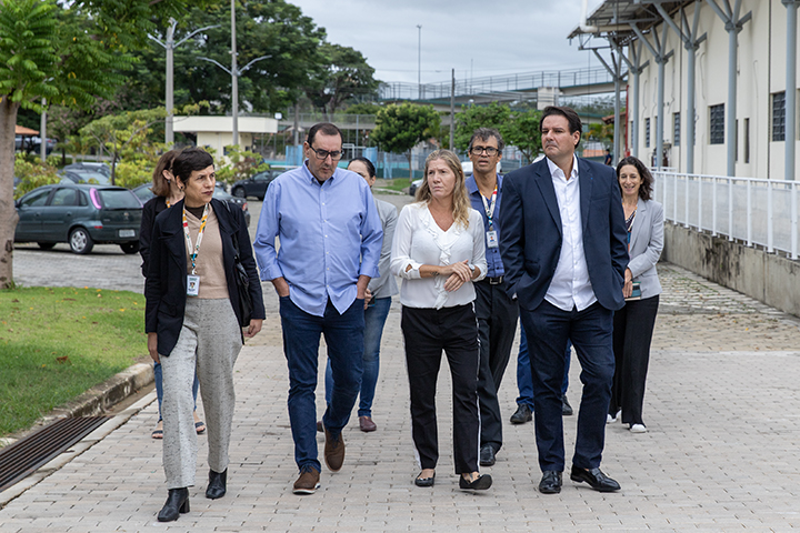 A imagem mostra um grupo de pessoas caminhando por uma área externa pavimentada, ao lado de um prédio institucional. Os integrantes estão em trajes formais e semi-formais, aparentando estar em uma visita técnica ou institucional. O ambiente é arborizado e bem cuidado, com árvores, carros estacionados e prédios ao fundo.
