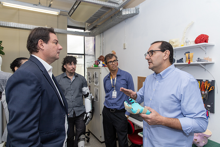 A imagem mostra um grupo de homens em um ambiente de laboratório ou oficina tecnológica. Um deles, vestindo camisa azul clara, está explicando algo enquanto segura uma prótese ou dispositivo robótico. Os demais observam atentamente, e um dos presentes usa uma prótese de braço. Ao fundo, há prateleiras com modelos anatômicos, ferramentas e peças de impressão 3D, indicando que o local está relacionado a pesquisa, robótica ou reabilitação. A cena sugere uma visita institucional ou técnica a um centro de inovação.