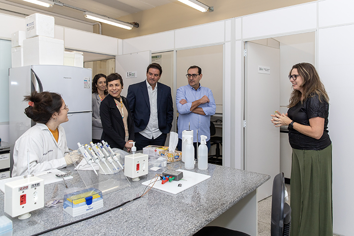 A imagem mostra uma visita a um laboratório científico. Uma pesquisadora com jaleco está sentada à esquerda, em uma bancada com micropipetas e outros materiais de laboratório. Um grupo de cinco visitantes, vestidos com roupas formais e semi-formais, observa e interage com ela, aparentando interesse. Uma mulher de óculos, à direita, parece estar apresentando ou explicando o funcionamento do laboratório. O ambiente é limpo, organizado e típico de pesquisa científica, com divisórias, equipamentos e iluminação fluorescente.