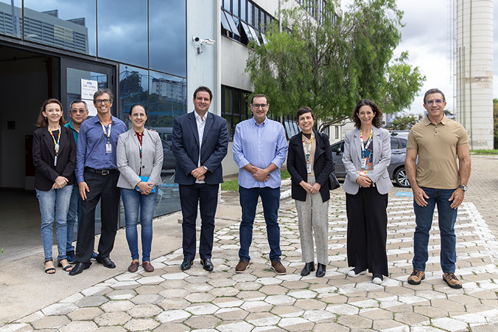A imagem mostra um grupo de dez pessoas posando para uma foto em frente a um prédio corporativo. Elas estão organizadas em uma fileira, a maioria vestindo roupas formais ou semi-formais. O ambiente parece ser institucional, com fachada de vidro, calçada de blocos intertravados e árvores ao fundo.